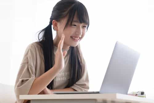 中学生の女の子が笑顔でオンライン個別指導を受け、パソコンに向かって手を挙げている写真