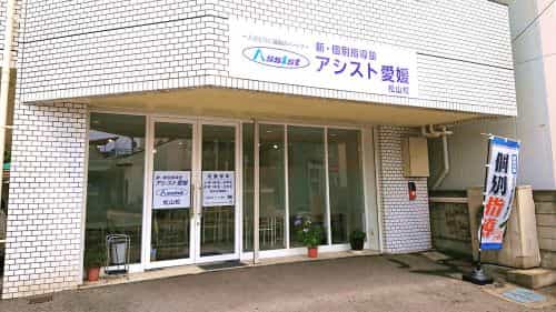 白い建物に新個別指導塾アシスト愛媛松山校の看板と個別指導の旗が写っている写真